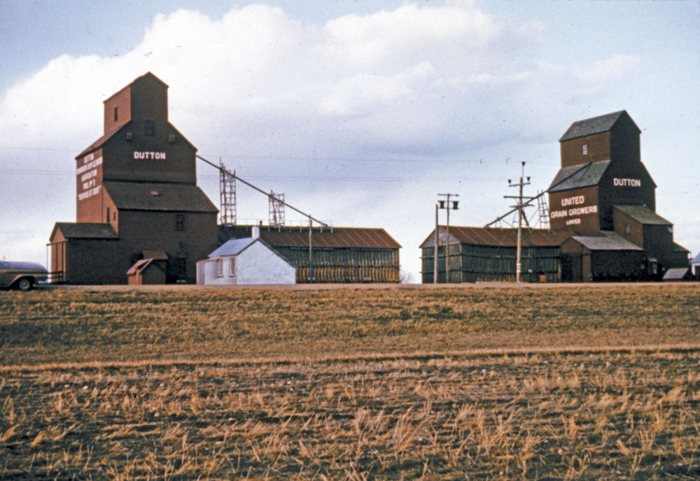 There were two chocolate-brown elevators at the Dutton Siding, on the CNR Togo Subdivision near Gilbert Plains, when this photo was taken in 1964. The UGG elevator at right had been built in 1920 and its annex was added in 1940. The left elevator was built by Paterson Grain before being sold in 1925 to Manitoba Pool. It was traded to UGG in late 1971. Between October and December 1976, both elevators were closed and later demolished.