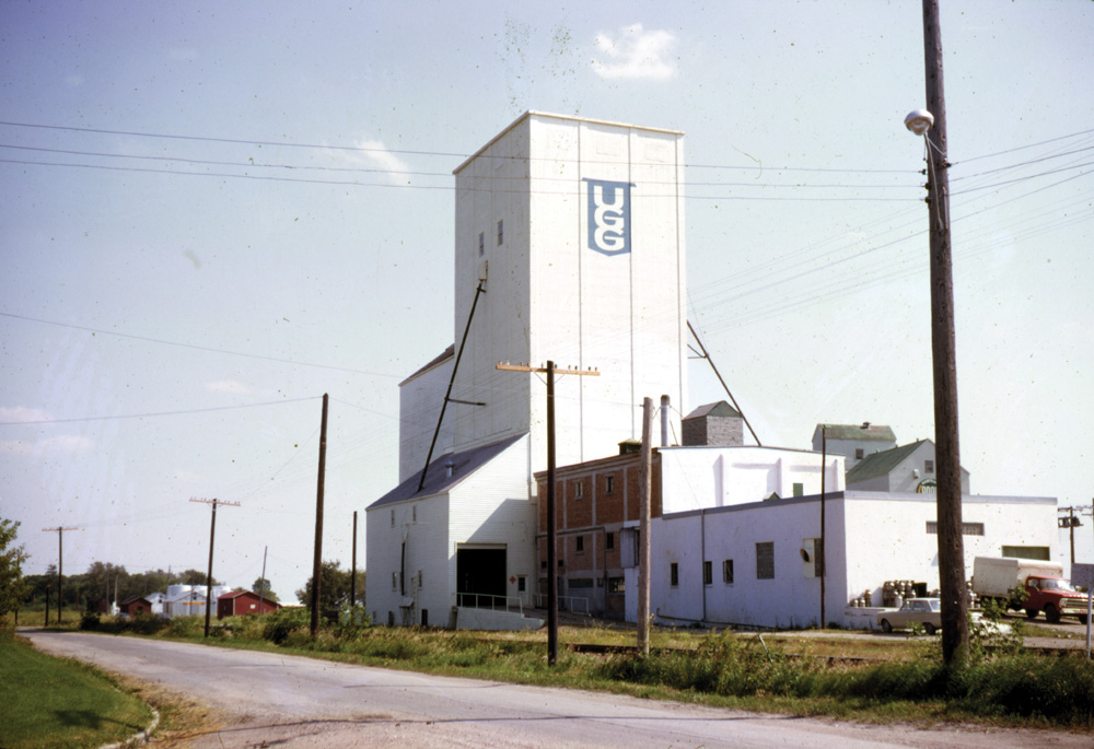 A grain elevator at Gladstone, shown here in 1969, held the double distinction of being the oldest country elevator in Manitoba made of concrete, and the first one to be demolished. Built in 1918 as a grist mill for the Echo Flour Mills Company, the mill portion was badly damaged by fire in 1919 but its grain elevator was left intact. The former mill building was used as a creamery until 1974 while the elevator was leased successively by Paterson Grain (1920-26), Manitoba Pool (1926-32), and United Grain Growers (1932-40). In July 1940, UGG purchased it outright. The facility closed in 1997.