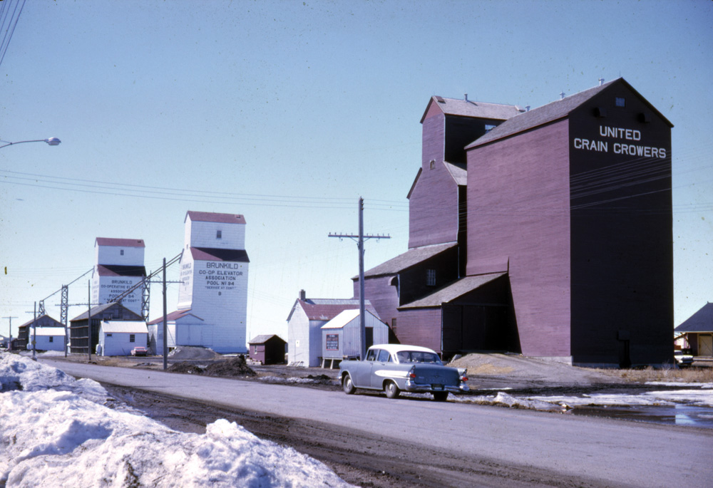 At the time of this 1962 photo, Brunkild had three grain elevators, one  operated by United Grain Growers and two by Manitoba Pool (A at left, B  in the middle). The UGG agent was G. A. Angus while the Pools were  managed by Gene Ferens (A) and Joseph Storey (B). Only the Pool A  elevator survives today, in use by BESCO Grain.