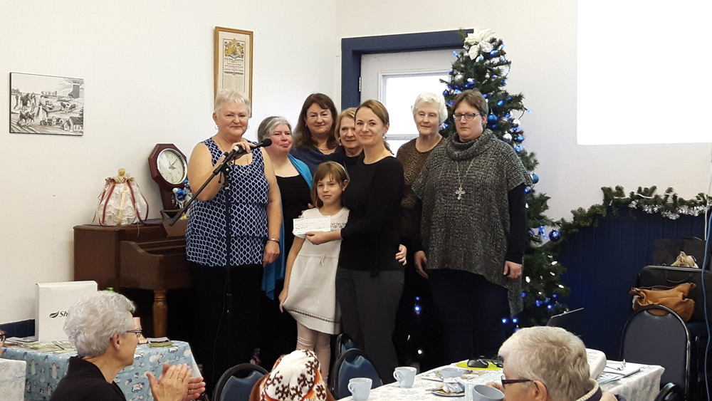 Bev Bennett (with microphone), back row (l to r), Julie McCahon, Janice Hunt, Dianna Airey, Wilma Bell, Wenda Best, present a cheque to Stacey Grocholsky and her daughter Kennedy.