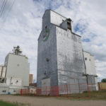 The history of ownership of this grain elevator, along Pacific Avenue in Brandon, is illustrated on its four sides. It was built around 1937 for the McCabe Brothers Grain Company, as shown by the name on its west  side. In 1965, the company erected six steel grain tanks and a feed-processing mill immediately east of the elevator. McCabe sold its elevators to United Grain Growers in early 1968 but this facility was excluded from the deal. Later that year, McCabe’s remaining assets were purchased by National Grain, named on its east side. The elevator (and the nearby mill) was bought in 1974 by Cargill, whose corporate signage is displayed on its north and south sides. At one time served by a spur line of the Canadian Pacific Railway that ran along its north side, there is no longer any rail service. Although slated for demolition in early 2016, it was still standing when this photo was taken in June 2016.