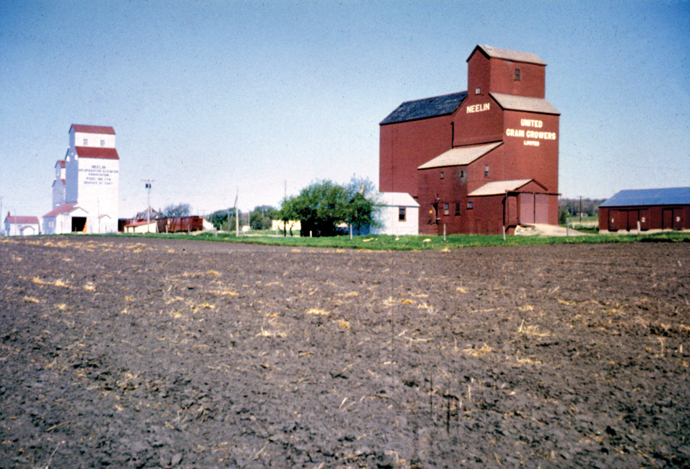 Around 1903, Western Canada Flour Mills built a 25,000-bushel elevator at Neelin, on the newly built CNR Wakopa Subdivision through the Rural Municipality of Argyle. In 1928, it was joined by a 30,000-bushel elevator constructed by United Grain Growers. By the time of this 1964 photo, the Western Canada elevator had been sold to Manitoba Pool, in 1940, and had been converted into an annex for a new elevator built in 1956. The mid-1950s also saw the addition of a 51,000-bushel crib annex beside the UGG elevator. Elevator and annex were traded to Manitoba Pool in 1973. Closed by December 1978, when the railway line was abandoned, the elevators have been removed from the site.