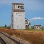 A railway track, overgrown with vegetation, runs beside an old elevator at Napinka. It was constructed in 1926 by the Ogilvie Milling Company. Its initial capacity of 35,000 bushels was expanded in 1957 with a 25,000-bushel annex. Taken over by Manitoba Pool in 1959, it became the B elevator at this point, used mostly for storing rye and oats. The Pool A elevator, with a 40,000-bushel capacity, was built in 1927 and expanded in 1951 with a 30,000-bushel annex. Later, Pool A was upgraded with a metric scale and larger driveway. The two elevators were operated by a single agent. The annexes were removed in the 1970s and the elevators were closed in July 1984, after efforts to sell them to another company failed. They were sold for use in private grain storage.