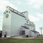 The former Manitoba Pool elevator at Oakville, seen here in a 1992 photo, was built between 1974 and 1975 to replace an elevator from 1928 destroyed by fire. An early example of the wooden composite type in which the elevator and annex are built as one solid crib, three steel tanks were added in 1990, increasing its capacity from 162,000 to 204,000 bushels. The facility continued to operate after the corporate merger that formed Agricore in 1998, but was closed temporarily in January 2001 as a cost-cutting measure then closed permanently in November 2001 after the Agricore merger with United Grain Growers. It is now used for private grain storage.