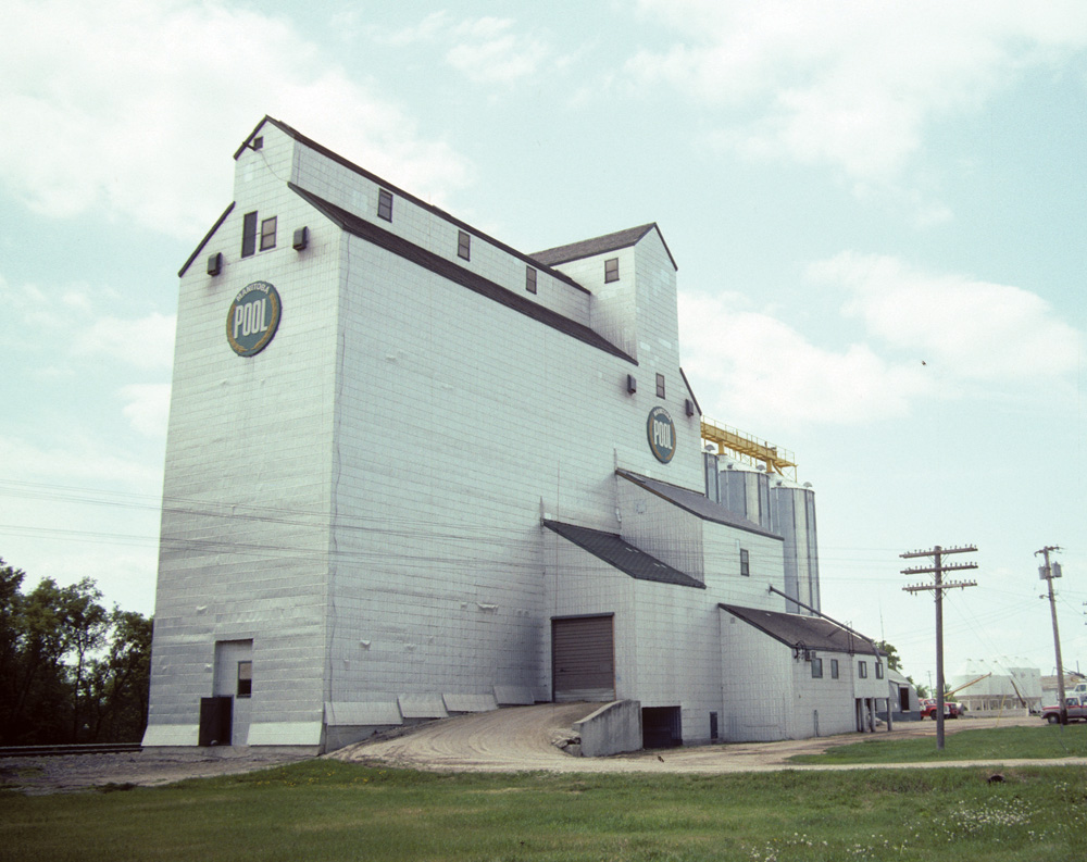 The former Manitoba Pool elevator at Oakville, seen here in a 1992 photo, was built between 1974 and 1975 to replace an elevator from 1928 destroyed by fire. An early example of the wooden composite type in which the elevator and annex are built as one solid crib, three steel tanks were added in 1990, increasing its capacity from 162,000 to 204,000 bushels. The facility continued to operate after the corporate merger that formed Agricore in 1998, but was closed temporarily in January 2001 as a cost-cutting measure then closed permanently in November 2001 after the Agricore merger with United Grain Growers. It is now used for private grain storage.