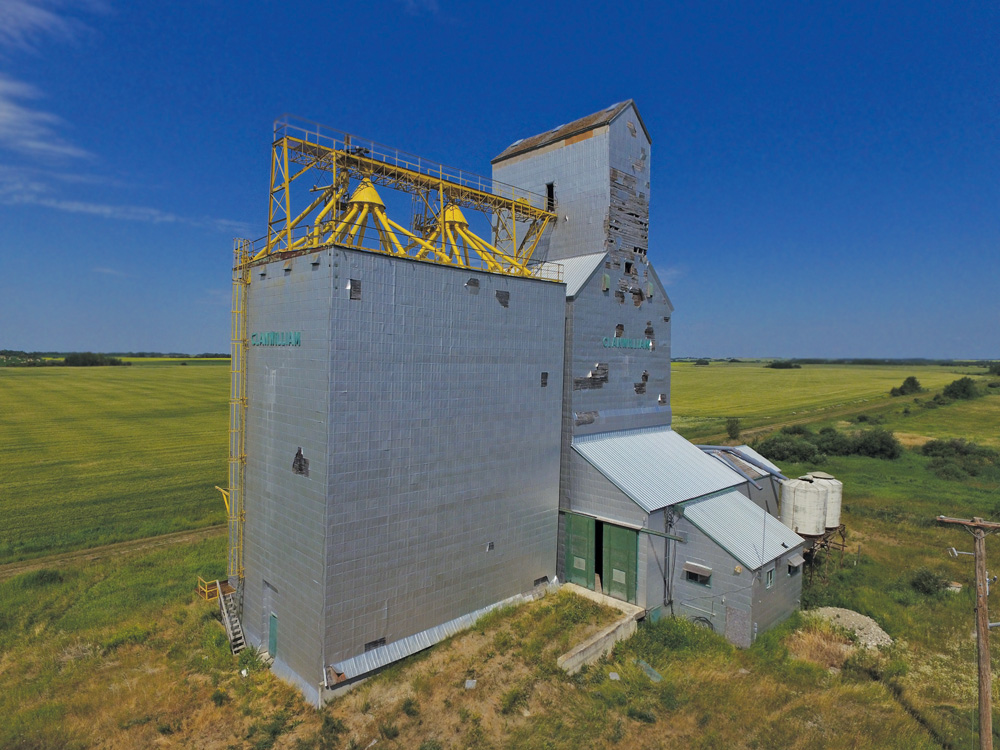 A 40,000-bushel grain elevator in Clanwilliam, in the RM of Minto-Odanah, was built in 1928 and operated by a co-operative association affiliated with Manitoba Pool. By the 1960s, it had a balloon annex on one side and an older, smaller elevator on the other. In 1982, the elevator was fully modernized, the balloon annex was removed, and a new crib annex replaced the old elevator. Closed in April 1996 when the CNR Rossburn Subdivision was abandoned, the facility has not been used for several years. As this recent photo shows, its metal cladding is damaged in several places and the exposed wood is deteriorating.