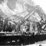 A steam shovel loading fill onto Manitoba and North Western flatcars. Steam shovels were capable of swinging the boom from side to side but the boom was fixed in position and only the bucket or dipper and the “dipper stick” could be raised and lowered. However, the dipper stick can be moved in and out. One can make out the rack bolted to the bottom of the dipper stick over the shoulder of the man in the bowler hat and vest. The large gear on the side of the boom worked a pinion which ran the rack back and forth. A small steam engine mounted on the boom worked this gear. The movable dipper stick made the steam shovel more effective. Other steam engines on the shovel swung the boom from side to side as required and worked the winch which raised and lowered the dipper.
