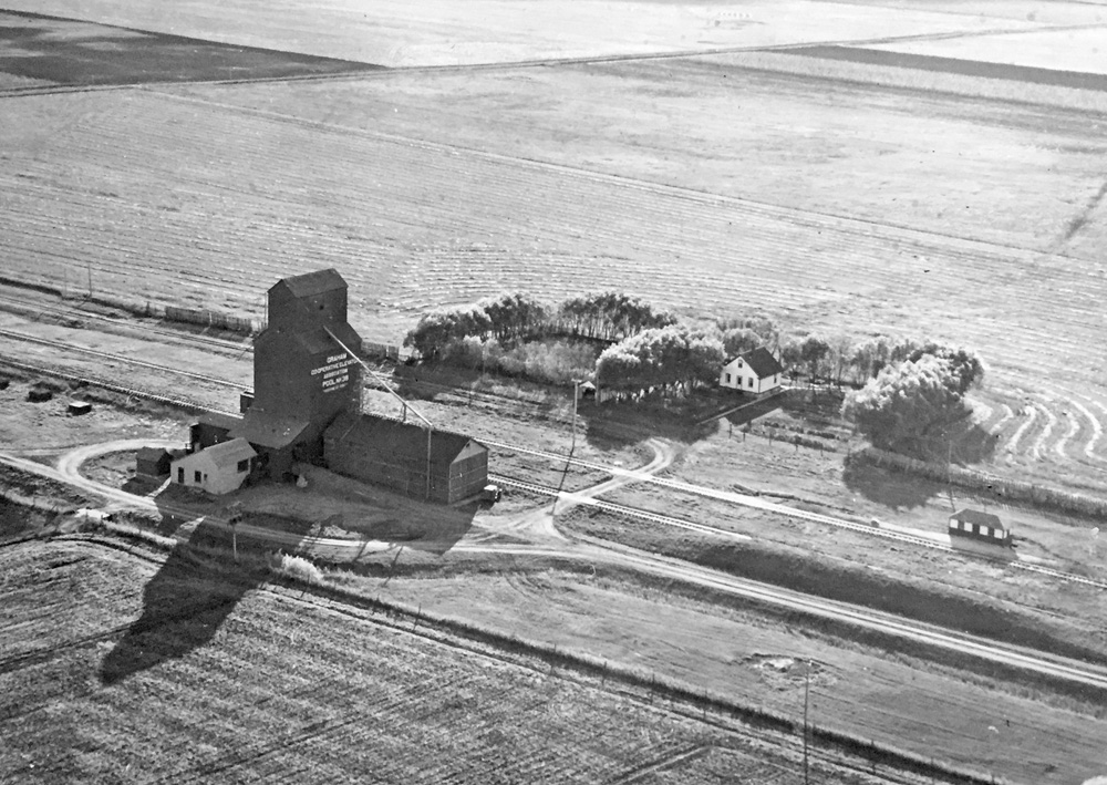 A 40,000-bushel wooden grain elevator at the railway siding of Graham, on the CPR Carman Subdivision in the Rural Municipality of Roland, was built in 1927 by Manitoba Pool Elevators. Closed in July 1971, the railway was abandoned in November 1975. No vestige of the railway tracks, elevator, agent’s residence, or access roads remain at the site in the middle of an agricultural field. This aerial view was provided by Doreen Smith whose husband Ralph was its agent from the mid-1940s to 1961.