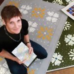Margruite Krahn, holding a copy of Resurfacing a field journal containing samples of floor designs, is a well-known Manitoba artist who lives in Neubergthal in southern Manitoba. The village is a national heritage site and was founded in 1876 by Mennonites who came from Prussia.