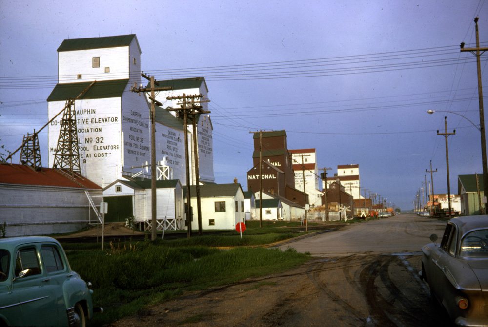 When this photo was taken in 1960, Dauphin’s “elevator row” along First Avenue had six members. At left was a Pool elevator dating from 1927 and labelled B when Pool A was built beside it in 1948. The brown National elevator in centre was built in 1952 and a preceding elevator was converted to an annex. Behind it was Pool D, acquired from Lake of the Woods Milling in 1959, that by 1962 had become United Grain Growers’ #2 elevator. At right was the original UGG elevator built around 1918, about the same time that another elevator, not visible behind UGG #1, was built by the Liberty Grain Company. It was sold to Searle Grain in 1929. All of them are now gone.