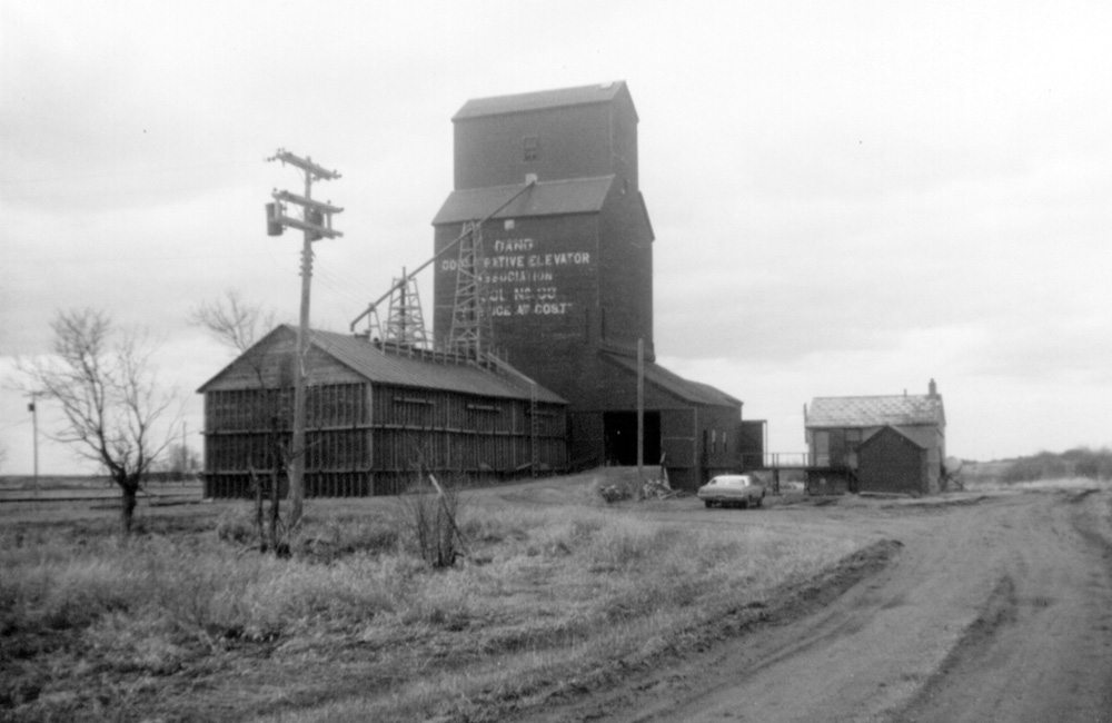 The railway siding of Dand, about 10 miles north of Deloraine, was established in 1913 after the CPR laid tracks in the area. The first grain elevator was built later that year by the Dominion Elevator Company. It was sold to the Canadian Consolidated Grain Company in 1928, the same year that Manitoba Pool built a 40,000-bushel elevator nearby. In addition to two grain elevators, at its height Dand consisted of a train station, general store, garage, church, school and teacherage, and a handful of houses. A 30,000-bushel balloon annex, visible in this photo from the 1970s, was built beside the Pool elevator in 1940. The Consolidated elevator was destroyed by fire in fall 1950. The Pool elevator closed in December 1974 and was later demolished. Today, a lonely church still stands at Dand.