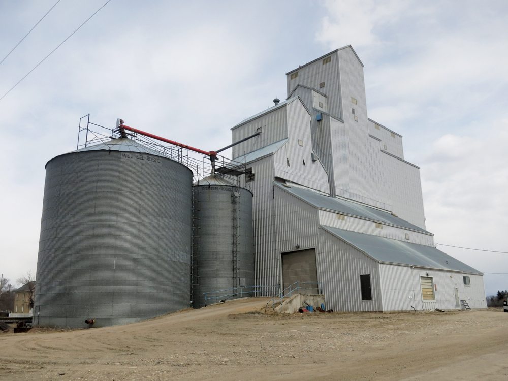 The oldest portion of this facility at Newdale, shown here in early 2017 between two steel bins and a larger elevator, was constructed by United Grain Growers in 1925. During the Second World War, in 1940 and 1941, two annexes were built beside it. One of the annexes was demolished in 1945 and the other was used until 1962. A third annex, constructed with the elevator in 1925, was removed in 1976. A thorough renovation occurred in 1966 and the steel bins (one with a capacity of 40,000 bushels and the other of 17,000 bushels) were erected on its west side in 1974. The larger elevator on the east side, with a capacity of 85,000 bushels, was built in 1983. Closed on July 31, 2000, the facility was sold to private ownership. The smaller elevator's foundation is cracked and, although its pit and leg are intact, it is no longer in use.