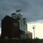 This photo from June 1981 shows a Manitoba Pool elevator at Franklin, northwest of Neepawa in the Rural Municipality of Rosedale, under renovation. The taller elevator was built in 1959 next to the shorter elevator from 1927. A balloon annex once stood to the right of the older elevator. The renovations in 1981 included a new office and larger driveshed, and installation of a metric scale. The crib annex at left, built in 1958, was moved here from Mentmore, 12 miles to the southeast. In 1991, the old elevator was demolished and replaced by three steel tanks. The 160,000-bushel elevator was closed in July 2000, following the opening by Agricore of a large, new facility at Minnedosa. The last elevator in the municipality was demolished in 2002.