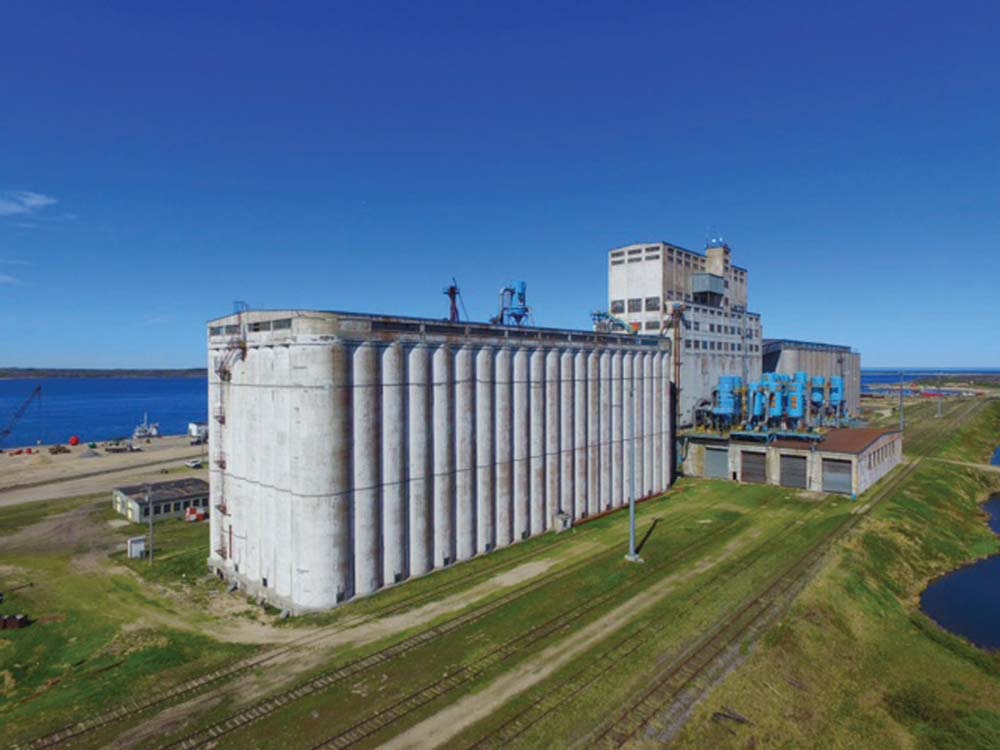 The Hudson Bay Railway reached Churchill in March 1929. One of the first priorities for construction was a terminal elevator for shipping grain to overseas markets. Designed by C. D. Howe & Company of Port Arthur (now Thunder Bay), it was erected by the Winnipeg firm of Carter Halls Aldinger between 1930 and 1931. A four-belt conveyor for loading up to four ships at once, at 20,000 bushels per hour, was completed in 1932. When done, the 2,500,000-bushel elevator was the second largest of its kind in the world. The 56 bins at its north (right) end, added between 1954 and 1955, doubled its capacity. Seen in this aerial view from June 2018, the elevator stands idle while the railway, damaged in May 2017, awaits repair.