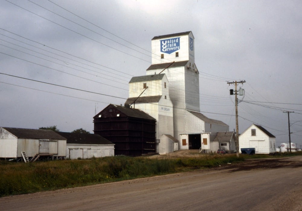 The first United Grain Growers elevator at McCreary, with a 30,000-bushel capacity, opened in 1917. Two balloon annexes were added to its east side, next to the driveway, around 1951 and its capacity rose to 85,000 bushels. Around 1961, one of the balloon annexes was moved to the south side and enlarged. In 1977, the old elevator was converted into an annex and a new elevator was built beside it. When this photo was taken in 1981, the original elevator and its single balloon annex were about to be demolished. Steel tanks on the south and north sides were added around 1995, increasing the total capacity to 141,000 bushels. Closed in 2001, the elevator was sold into private ownership. At the time of a 2017 visit, it appeared to be disused except by many pigeons. 