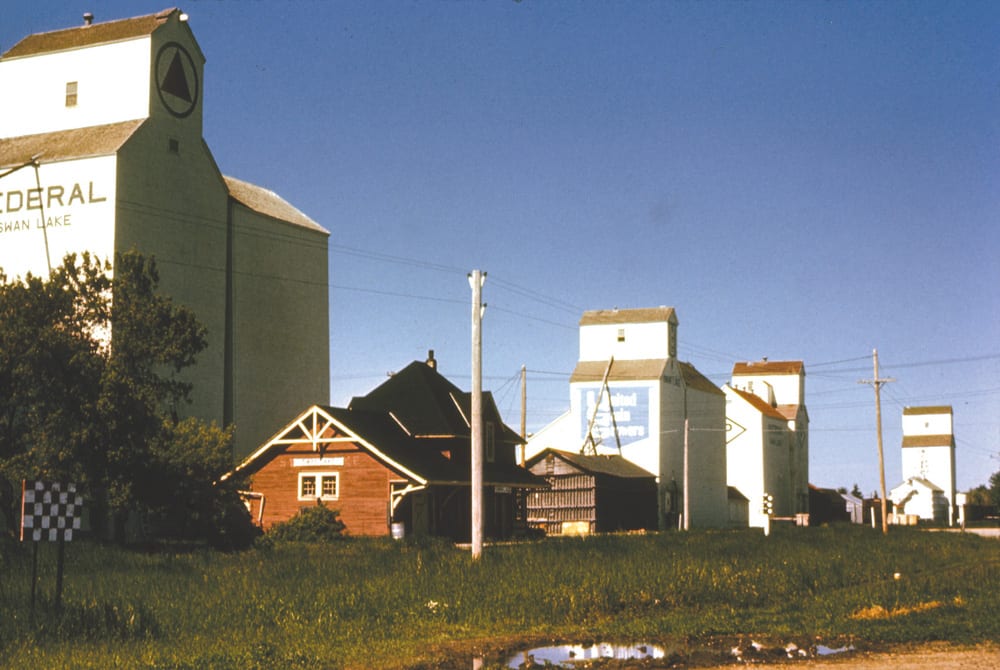 In this 1971 photo of Swan Lake, we see the local train station near four elevators, all of them gone today. From left to right (east to west), we see the Federal elevator built in 1954 that was sold to Manitoba Pool in 1960 then traded to UGG in 1971 (but the Federal name remained on it); then the UGG elevator built in 1925; then the Paterson elevator dating from 1924 with its crib annex (1953) and balloon annex (1940); and finally the 1928 Manitoba Pool elevator traded to UGG in 1974 and burned in 1988.