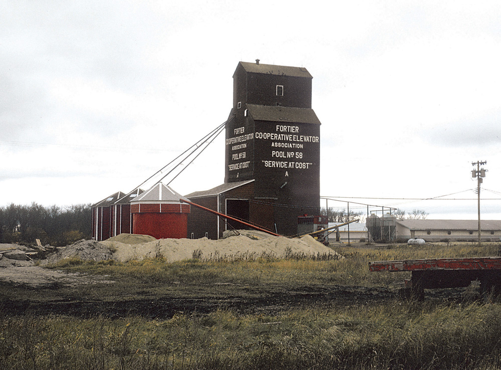 In 1965, the provincial government bought two elevators and annexes at Fortier, about 24 kilometres (15 miles) southeast of Portage la Prairie, from Manitoba Pool. The “A” elevator had been built by Pool in 1928 while the “B” elevator dated from 1913 and had been bought from Ogilvie Flour Mills in 1959. The government used the abandoned right-of-way for the CN railway line beside the elevators to expand the Trans-Canada Highway from two to four lanes. By the time of this 1973 photo, the former A elevator had been moved to a nearby Hutterite colony.