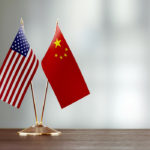 American and Chinese flag pair on desk over defocused background. Horizontal composition with copy space and selective focus.