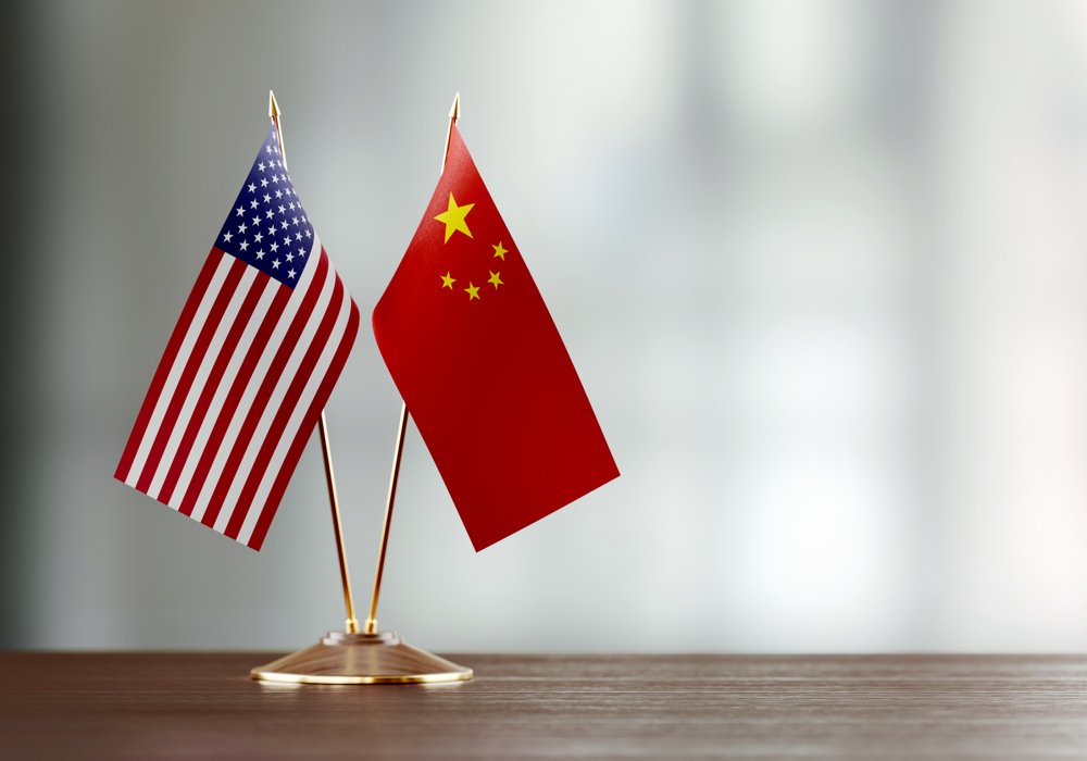 American and Chinese flag pair on desk over defocused background. Horizontal composition with copy space and selective focus.