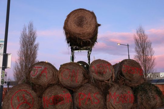 A hay bale is moved by a tractor over a pile to block a road (not the road linking France and Spain) near Castelnaudary, in the south of France, January 23, 2024. The text on the hay bales reads: “JA” referring to “Jeunes Agriculteurs” (Young Farmers) and “We just want to feed, not die”. Photo: Reuters/Nacho Doce
