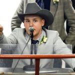 A man in a light suit jacket and charcoal coloured cowboy hat chants into the microphone while pointing out bids