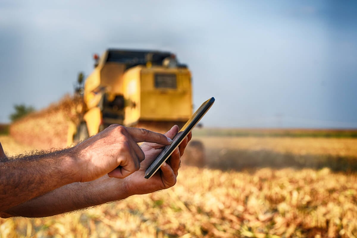 A farmer holds a cell phone with a combine harvesting crop in the background.