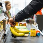 A person putting some bananas and other groceries on the conveyor at a store checkout.