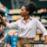 A thoughtful woman shops in a supermarket, browsing through products on the shelves. She seems to be making a careful decision about her purchase.