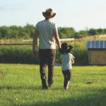 Father and child walk across a vibrant green field on a sunny day, evoking feelings of connection, nature, and simplicity, surrounded by rural scenery perfect for family-themed concepts and peaceful environments.