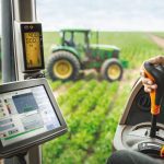 View from inside a tractor cab with a hand on the gear shift and a digital display screen showing field data during precision agriculture operations. Photo: Bayer AG
