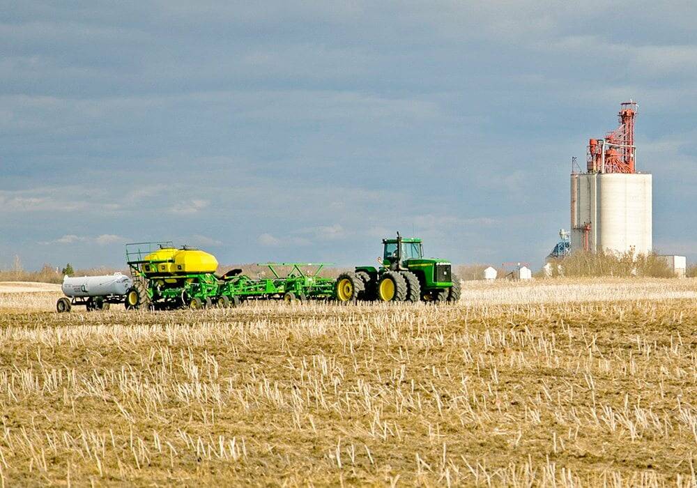 Tractor pulling a fertilizer applicator and anhydrous ammonia tank across a stubble field on the Prairies, with a grain elevator in the background. Photo: file