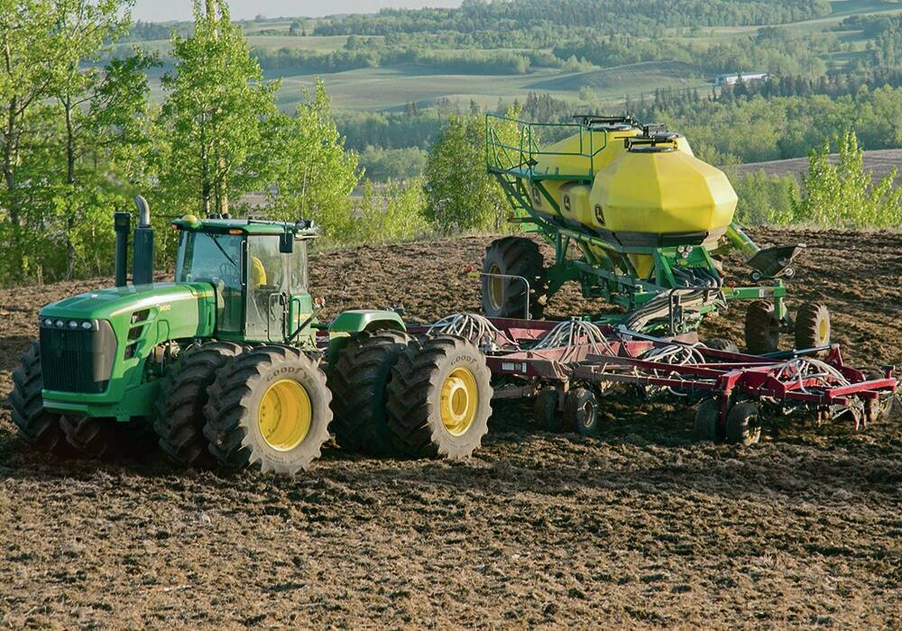 A green tractor pulls a red air seeder and green and yellow cultivator across a worked field with rolling green hills in the background as Manitoba input costs for fertilizer and fuel squeeze producers heading into 2026. Photo: Robin Booker