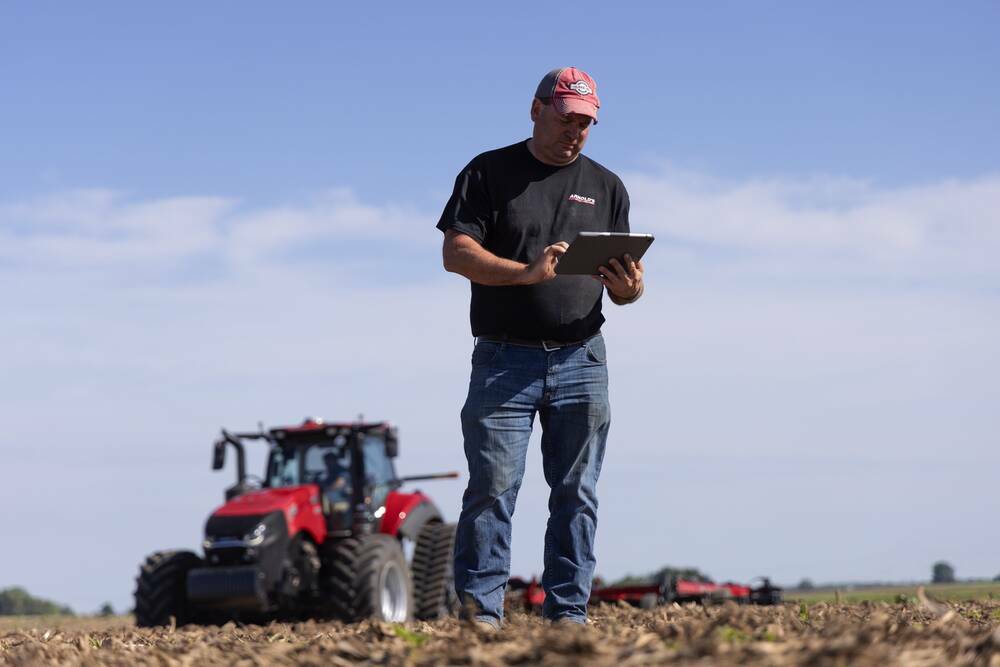 A farmer stands in a bare field checking a tablet with a tractor and seeding equipment behind him as digital tools reshape on-farm agronomy. Photo: CNH