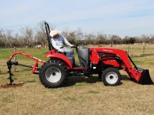 Operator using a red Mahindra 1533 compact tractor with front loader and post-hole auger on a rural property. Photo: PRNewsFoto/Mahindra.