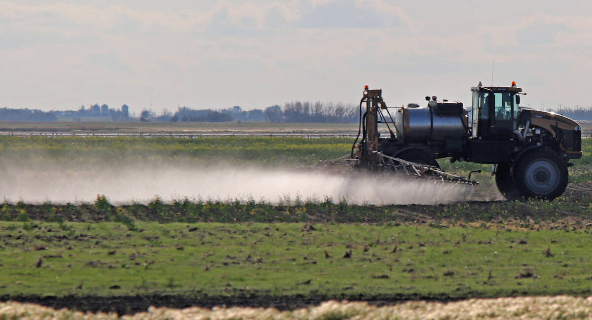 A self-propelled sprayer applies herbicide across a flat Manitoba field in early season, with weed patches visible on bare ground. Photo: Robin Booker.