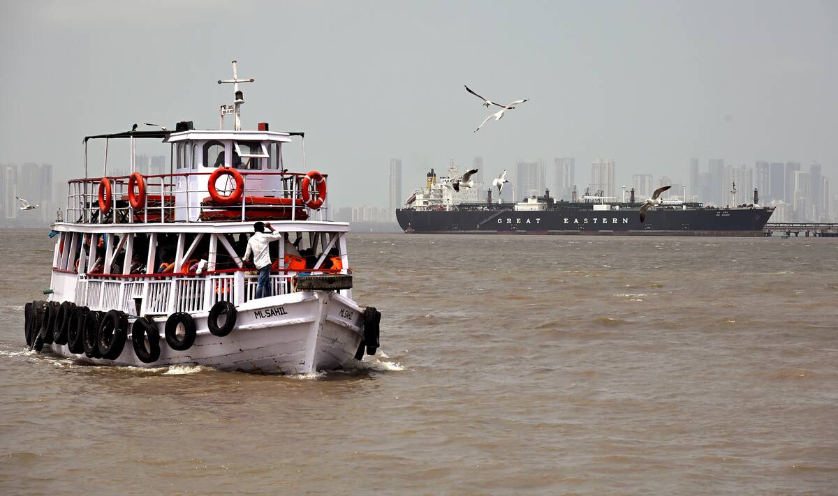 A boat in the foreground with a large tanker ship anchored in hazy waters at Mumbai port, connecting Gulf region geopolitics to Canadian fertilizer prices. Photo: Ashish Vaishnav / SOPA Images via Reuters Connect.