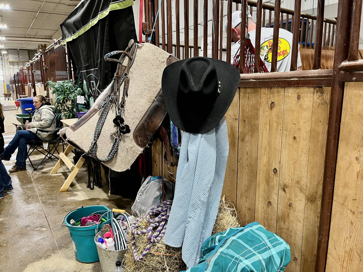 A cowboy hat, saddle blanket and tack hanging on a wooden stall divider in a barn at the Royal Manitoba Winter Fair, with hay bales and equipment visible.Photo: Alexis Stockford