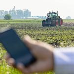 A hand holding a smartphone in the foreground with a Fendt tractor working a field in the background, illustrating the OutRun autonomous tillage system's remote operation capability. Photo: Agco/Fendt