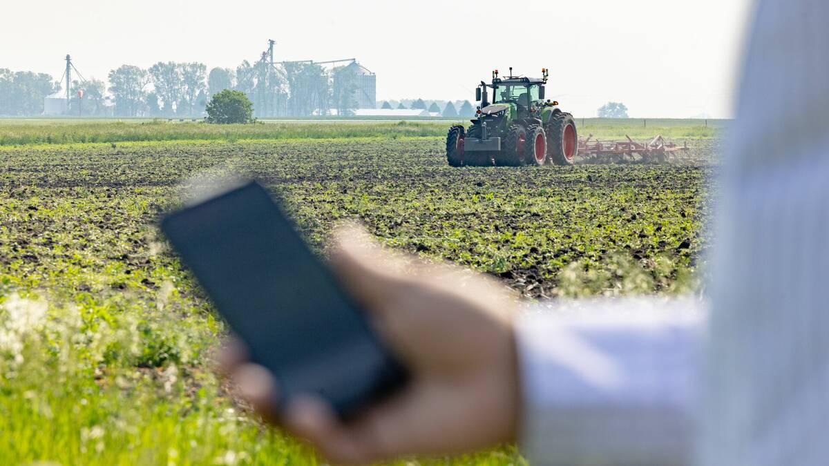 A hand holding a smartphone in the foreground with a Fendt tractor working a field in the background, illustrating the OutRun autonomous tillage system's remote operation capability. Photo: Agco/Fendt