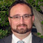 Headshot of Cam Dahl, general manager of Manitoba Pork, wearing glasses and a dark suit jacket, photographed outdoors with green foliage in the background. Photo: file.