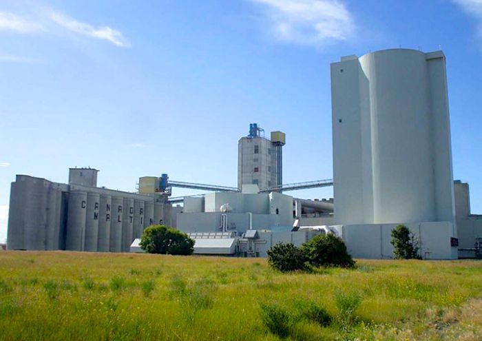 The Canada Malting plant in Calgary, with grey concrete silos and white processing towers set against a blue sky, viewed across a grassy field. Photo: file