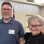 Jon Whetter wearing a name tag and Dianne Kohler in glasses standing side by side at an indoor event, with a Manitoba flag visible in the background. Photo: Gord Gilmour