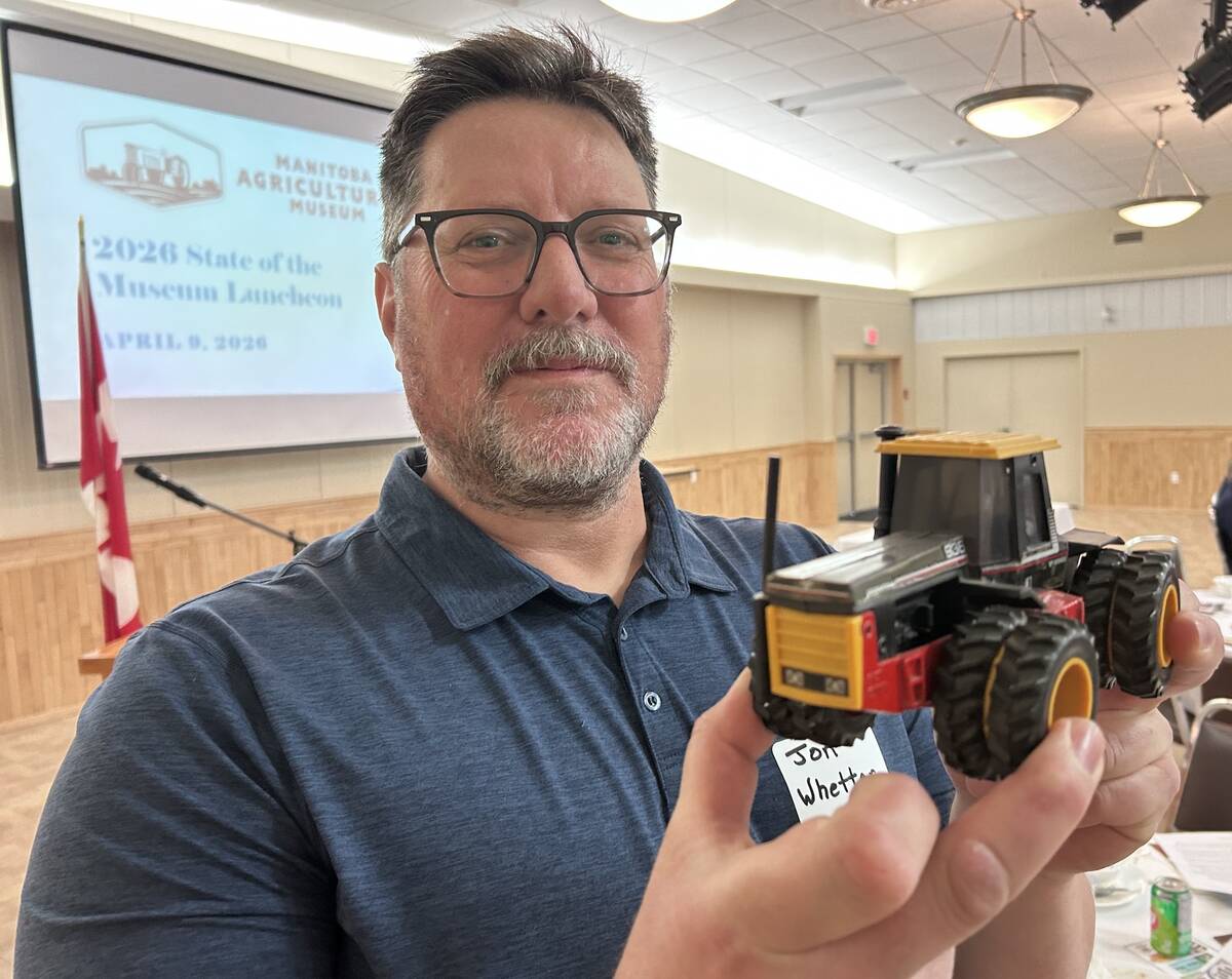 Jon Whetter holding a yellow and red Versatile model tractor at a luncheon, with a Manitoba Agricultural Museum presentation slide visible on screen behind him. Photo: Gord Gilmour