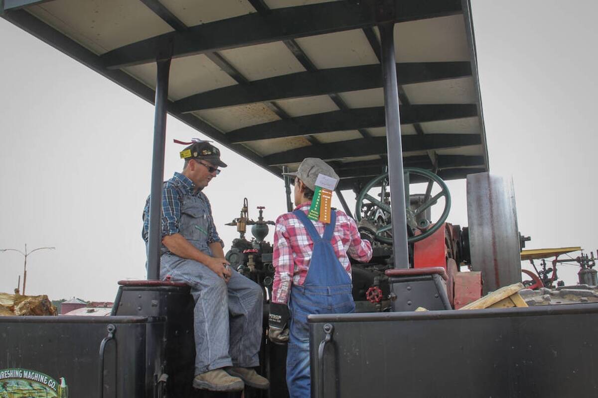 An older man in a ball cap and a young boy in overalls and a plaid shirt standing on a vintage steam engine at an outdoor agricultural event. Photo: Geralyn Wichers