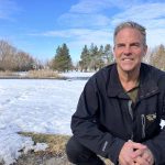 Daniel Bezte sitting outdoors on his snow-covered property with melting ice, grass, and trees in the background under a blue sky. Photo: Greg Berg.