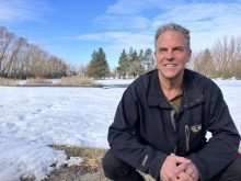 Daniel Bezte sitting outdoors on his snow-covered property with melting ice, grass, and trees in the background under a blue sky. Photo: Greg Berg.